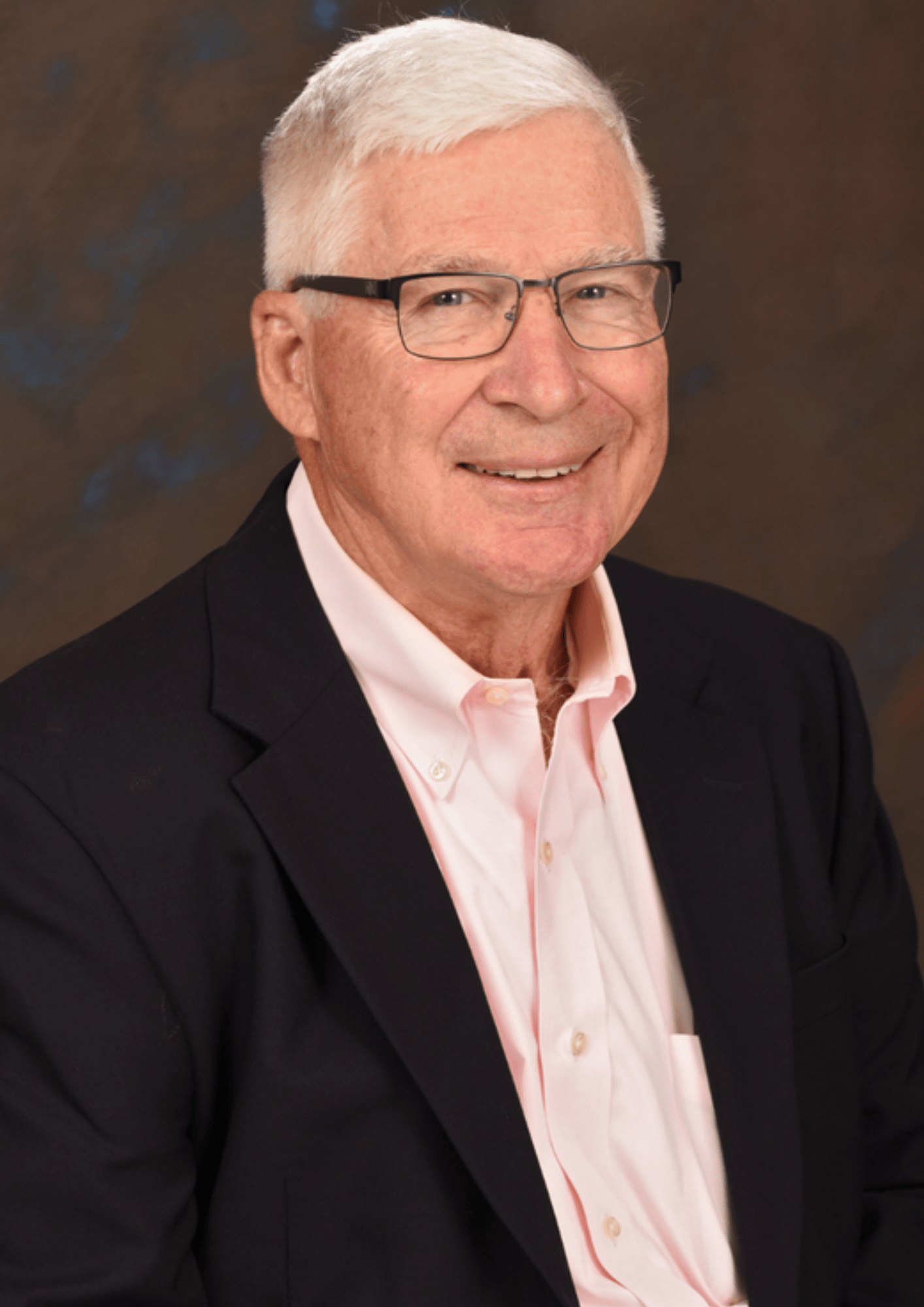 An older man wearing glasses and a pink shirt, smiling gently while seated in a well-lit room.